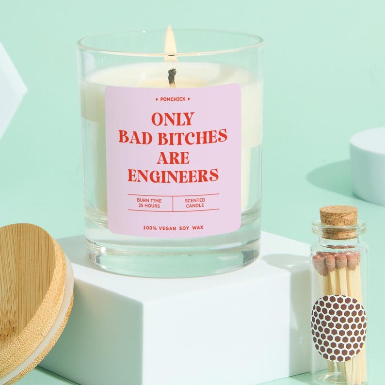 May include: A lit scented candle in a clear glass jar with a pink label that reads "ONLY BAD BITCHES ARE ENGINEERS." The candle is made of 100% vegan soy wax and has a burn time of 25 hours. A small glass bottle of matches is next to the candle.