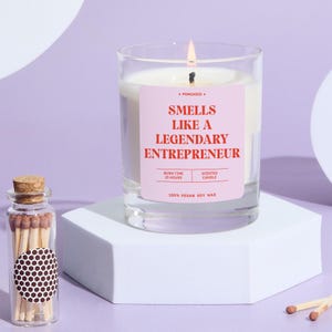 May include: A lit scented candle in a clear glass jar with a pink label that reads "Smells Like a Legendary Entrepreneur." The candle sits on a white block, with a small glass jar of matches to the left. The candle is made of 100% vegan soy wax.