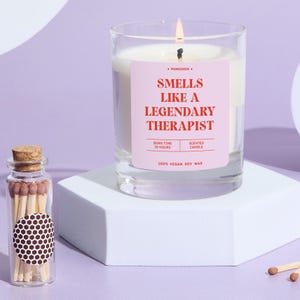 May include: A lit scented candle in a clear glass jar with a pink label that reads "Smells Like a Legendary Therapist." A small glass bottle of matches sits nearby. The candle is made of 100% vegan soy wax and has a 25-hour burn time.