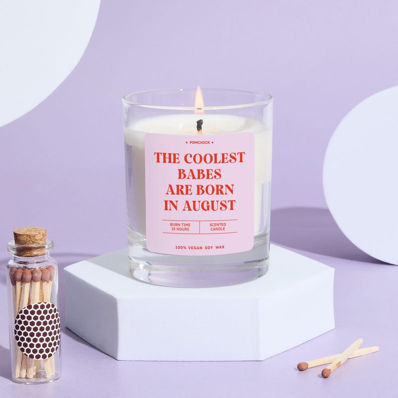 May include: A lit scented candle in a clear glass jar with a pink label that reads "THE COOLEST BABES ARE BORN IN AUGUST." A small glass bottle of matches and two matches are also visible. The candle is on a white geometric stand.