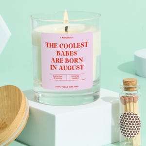 May include: A lit scented candle in a clear glass jar with a pink label that reads "THE COOLEST BABES ARE BORN IN AUGUST". The candle is made of 100% vegan soy wax and has a burn time of 25 hours. A small glass jar of matches is next to the candle.