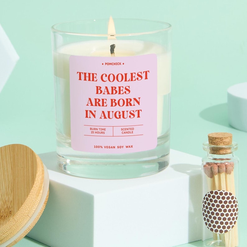 May include: A lit scented candle in a clear glass jar with a pink label that reads "THE COOLEST BABES ARE BORN IN AUGUST". The candle is made of 100% vegan soy wax and has a burn time of 25 hours. A small glass jar of matches is next to the candle.