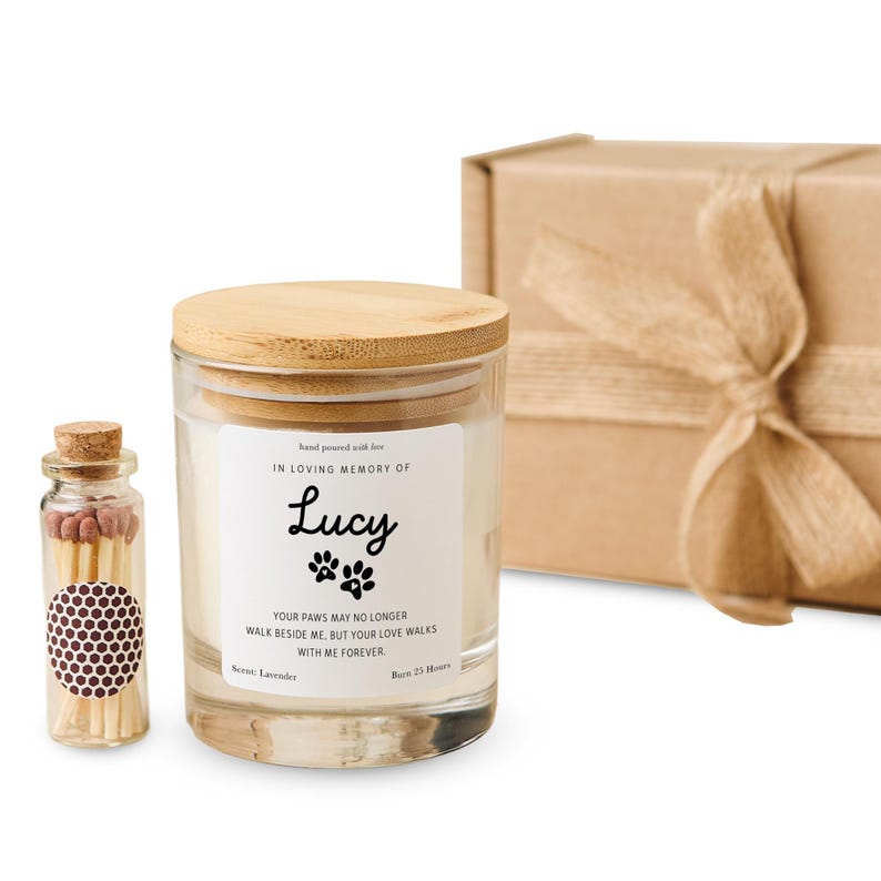 Pode incluir: Um frasco de vela de vidro com tampa de madeira, uma pequena garrafa de vidro de f&oacute;sforos e uma caixa de presente embrulhada. A vela tem um r&oacute;tulo que diz "IN LOVING MEMORY OF Lucy" com pegadas e uma mensagem. A vela tem aroma de lavanda e queima por 25 horas.