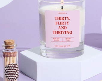 Może przedstawiać: Zapachowa świeca w szklanym słoiku z różową etykietą z napisem "THIRTY, FLIRTY AND THRIVING". Obok świecy stoi mały szklany słoik z zapałkami. Świeca wykonana jest w 100% z wegańskiego wosku sojowego i ma czas palenia 25 godzin.