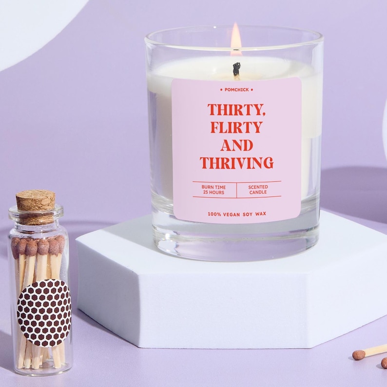 May include: A lit scented candle in a clear glass jar with a pink label that reads "THIRTY, FLIRTY AND THRIVING." A small glass jar of matches sits next to the candle. The candle is made with 100% vegan soy wax and has a burn time of 25 hours.