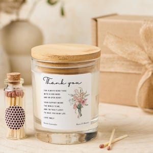 May include: A glass candle with a wooden lid and a label that says "Thank you." The label features a floral design and text. A small glass bottle of matches is to the left. The candle is lavender scented.