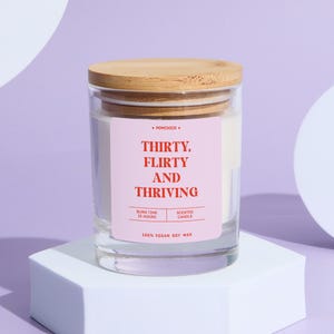 May include: A clear glass candle jar with a wooden lid. The label reads "THIRTY, FLIRTY AND THRIVING" in red text on a pink background. The candle is filled with white wax and is labeled as 100% vegan soy wax.