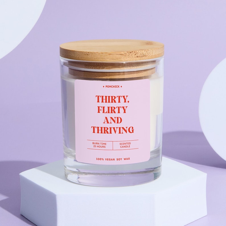 May include: A clear glass candle jar with a wooden lid. The label reads "THIRTY, FLIRTY AND THRIVING" in red text on a pink background. The candle is filled with white wax and is labeled as 100% vegan soy wax.
