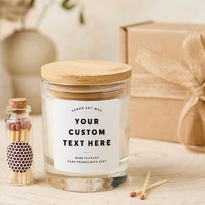Può includere: Un barattolo di candela in vetro trasparente con coperchio in legno ed etichetta bianca con la scritta "YOUR CUSTOM TEXT HERE". Nella foto sono presenti anche una piccola bottiglia di vetro con fiammiferi e una confezione regalo. La candela è fatta di cera di soia vegana.