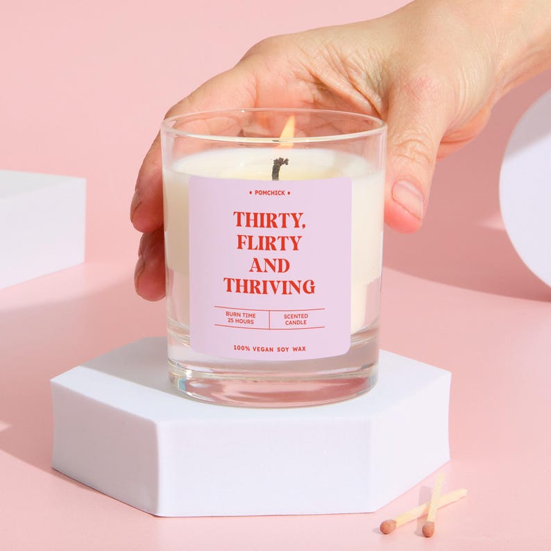 May include: A lit scented candle in a clear glass jar with the text "THIRTY, FLIRTY AND THRIVING" on a pink label. The candle is on a white hexagonal stand, with matches in the foreground. The candle is made of 100% vegan soy wax.