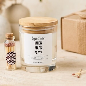 May include: A glass candle jar with a wooden lid and the text "light me WHEN MARK FARTS". A small glass bottle of matches and a cardboard box are also present. The candle is lavender scented and has a 25-hour burn time.