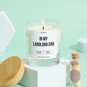 May include: A lit scented candle in a clear glass jar with the text "IN MY LANDLORD ERA" on a white label. The candle is on a white cube, with a wooden lid and a small glass bottle of matches nearby. The candle is made of 100% vegan soy wax.