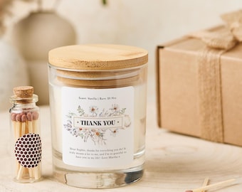 May include: A clear glass candle jar with a wooden lid, labeled with "THANK YOU" and floral accents. A small glass bottle of matches and a wrapped gift box are also present. The candle scent is vanilla and burns for 25 hours.