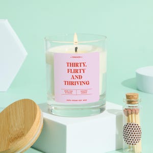 May include: A lit scented candle in a clear glass jar with a pink label that reads "THIRTY, FLIRTY AND THRIVING." The candle sits on a white cube, with a wooden lid and a small glass bottle of matches nearby.