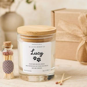 Pode incluir: Um frasco de vela de vidro transparente com tampa de madeira, rotulado "IN LOVING MEMORY OF Lucy" com pegadas de patas. A vela &eacute; acompanhada por um pequeno frasco de vidro com f&oacute;sforos e uma caixa de presente. Fragr&acirc;ncia de lavanda, tempo de queima 23 horas.