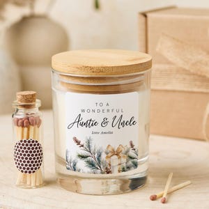 May include: A glass candle with a wooden lid and a label that reads "To a Wonderful Auntie & Uncle". The label features a gift box and pine branches. A small glass bottle of matches is next to the candle.
