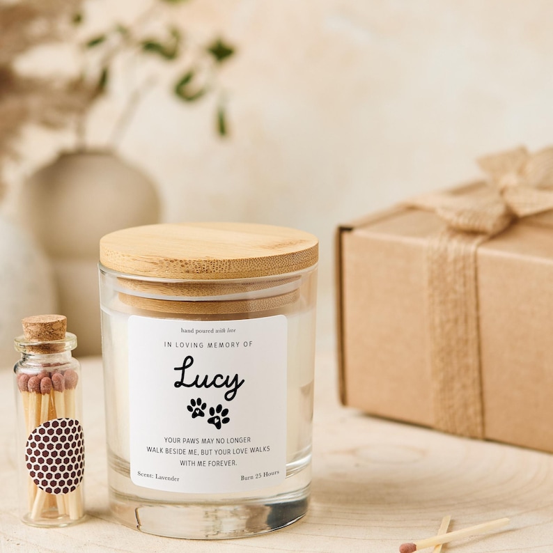 Pode incluir: Um frasco de vela de vidro transparente com tampa de madeira, rotulado "In Loving Memory of Lucy" com pegadas de patas. Uma pequena garrafa de vidro de f&oacute;sforos e uma caixa de presente embrulhada tamb&eacute;m est&atilde;o presentes. A vela &eacute; perfumada com lavanda e queima por 23 horas.
