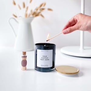 以下が含まれることがあります： 白いラベルに「YOUR CUSTOM TEXT HERE」と「Hand poured with love, Vegan soy wax candle」と書かれた黒いキャンドル。キャンドルに火をつけるためにマッチが燃やされています。画像には、マッチの入った小さなガラス瓶も写っています。