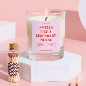 Könnte beinhalten: Eine brennende Duftkerze in einem transparenten Glas mit einem rosa Etikett mit der Aufschrift "Smells Like a Legendary Nurse". Eine kleine Glasflasche mit Streichhölzern befindet sich neben der Kerze. Die Kerze besteht aus 100 % veganem Sojawachs.