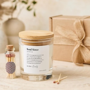 Puede incluir: Un tarro de vela de vidrio transparente con tapa de madera y una etiqueta que dice "Soul Sister". También hay una pequeña botella de vidrio con cerillas y una caja de regalo envuelta. La vela tiene aroma a lavanda.