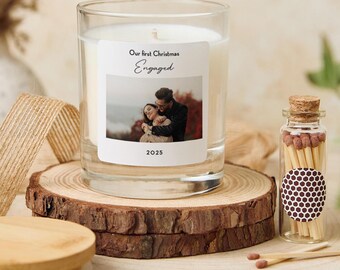May include: A clear glass candle with a white label that reads "Our first Christmas Engaged 2025" and features a photo of a couple. The candle sits on a wooden slice with a small glass jar of matches.