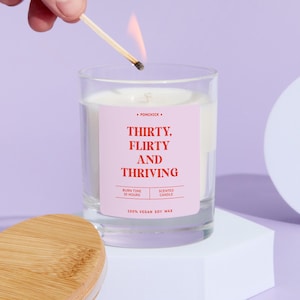 May include: A lit candle in a clear glass jar with a pink label. The label reads "THIRTY, FLIRTY AND THRIVING". The candle is on a white surface, with a wooden lid and a match lighting the wick. The candle is made of 100% vegan soy wax.