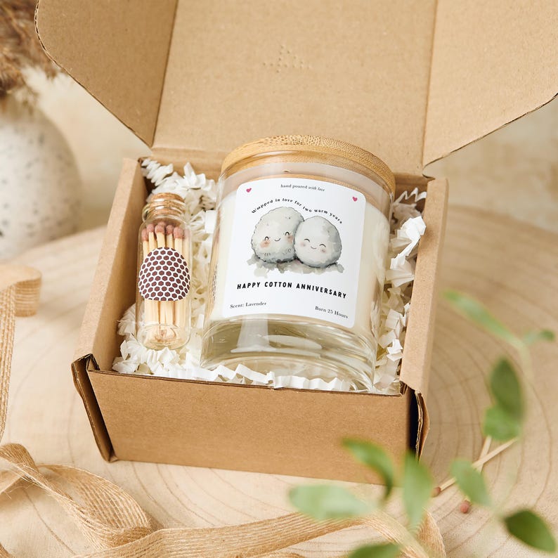 May include: A gift box containing a candle and matches. The candle has a wooden lid and a label with the text "Happy Cotton Anniversary" and a drawing of two smiling cotton balls. The match bottle has a brown and white pattern.