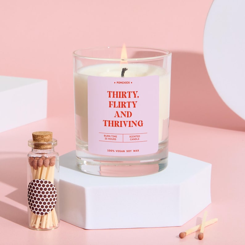 May include: A lit scented candle in a clear glass jar with a pink label that reads "THIRTY, FLIRTY AND THRIVING." A small glass bottle of matches and two matches are next to the candle. The candle is made of 100% vegan soy wax.