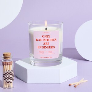 May include: A lit scented candle in a clear glass jar with a pink label that reads "ONLY BAD BITCHES ARE ENGINEERS." A small glass bottle of matches sits nearby. The candle is on a white geometric stand.