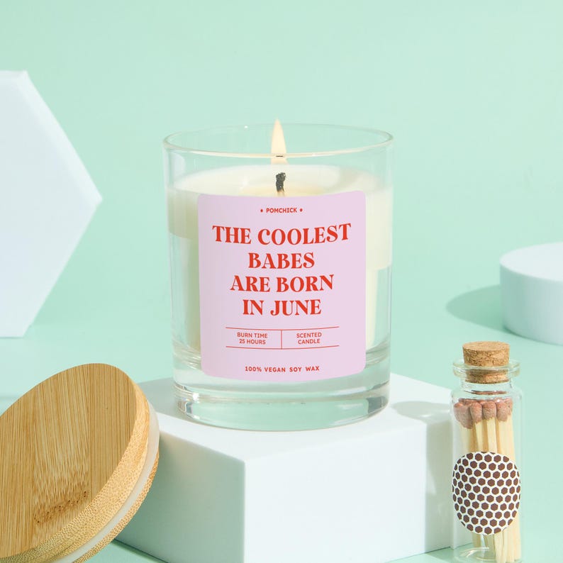 May include: A clear glass candle with a pink label that reads "THE COOLEST BABES ARE BORN IN JUNE." The candle is lit, and a small glass bottle of matches sits nearby. The candle is made of 100% vegan soy wax and has a burn time of 25 hours.