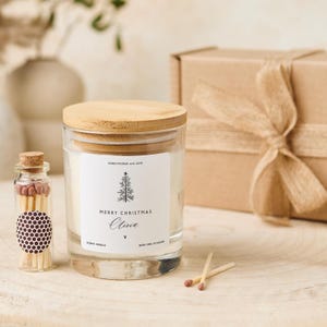 Può includere: Un barattolo di candela in vetro trasparente con coperchio in legno, etichettato "Merry Christmas Olivia" con una grafica di albero di Natale. Sono presenti anche una piccola bottiglia di vetro di fiammiferi e una confezione regalo avvolta. La candela è alla vaniglia e ha un tempo di combustione di 24 ore.