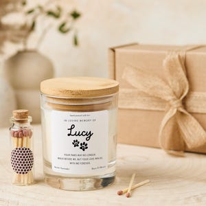 Pode incluir: Um frasco de vela de vidro com tampa de madeira, rotulado "IN LOVING MEMORY OF Lucy". O r&oacute;tulo inclui pegadas de patas e o texto "YOUR PAWS MAY NO LONGER WALK BESIDE ME, BUT YOUR LOVE WALKS WITH ME FOREVER". Uma pequena garrafa de vidro de f&oacute;sforos est&aacute; ao lado da vela.