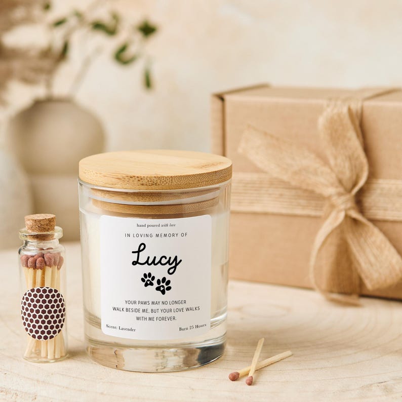 Pode incluir: Um frasco de vela de vidro com tampa de madeira, rotulado "IN LOVING MEMORY OF Lucy". O r&oacute;tulo inclui pegadas de patas e o texto "YOUR PAWS MAY NO LONGER WALK BESIDE ME, BUT YOUR LOVE WALKS WITH ME FOREVER". Uma pequena garrafa de vidro de f&oacute;sforos est&aacute; ao lado da vela.