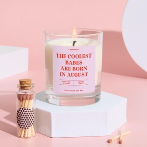 May include: A lit scented candle in a clear glass jar with a pink label that reads "THE COOLEST BABES ARE BORN IN AUGUST." A small glass bottle of matches and two loose matches are next to the candle. The candle is on a white geometric stand.