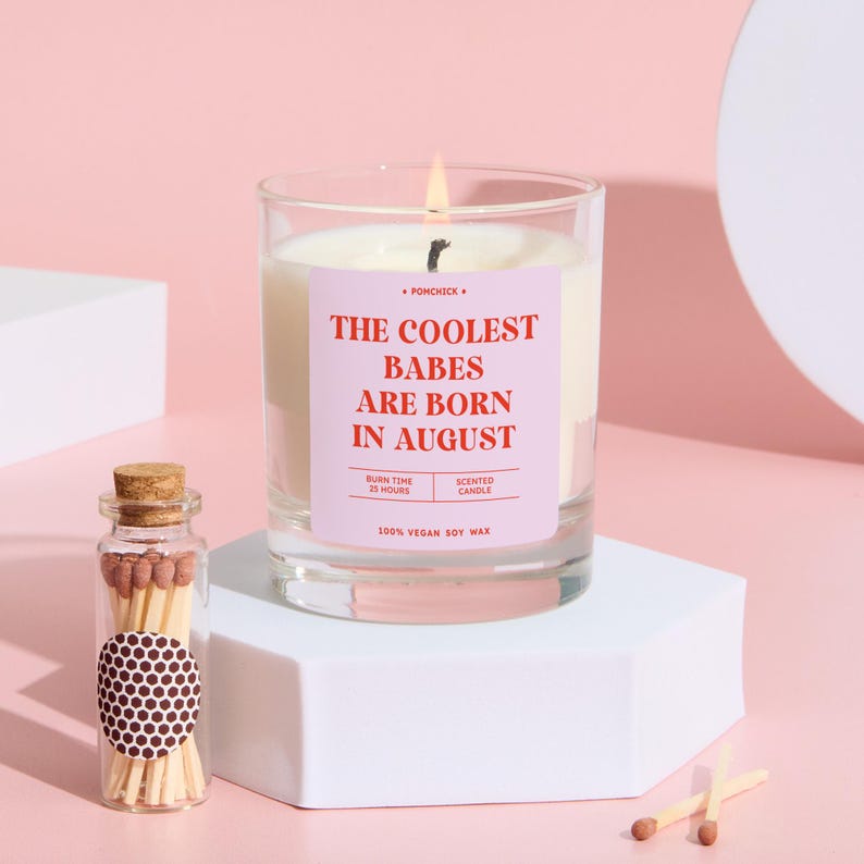 May include: A lit scented candle in a clear glass jar with a pink label that reads "THE COOLEST BABES ARE BORN IN AUGUST." A small glass bottle of matches and two loose matches are next to the candle. The candle is on a white geometric stand.