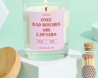 May include: A lit scented candle in a clear glass jar with a pink label that reads "ONLY BAD BITCHES ARE LAWYERS." The candle is on a white cube, with a small glass jar of matches to the right. The candle is made of 100% vegan soy wax.
