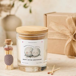 May include: A glass candle jar with a wooden lid, labeled "Happy Cotton Anniversary." The label features two cartoon cotton balls and the text "Wrapped in love for two warm years." A small glass bottle of matches and a gift box are also present.