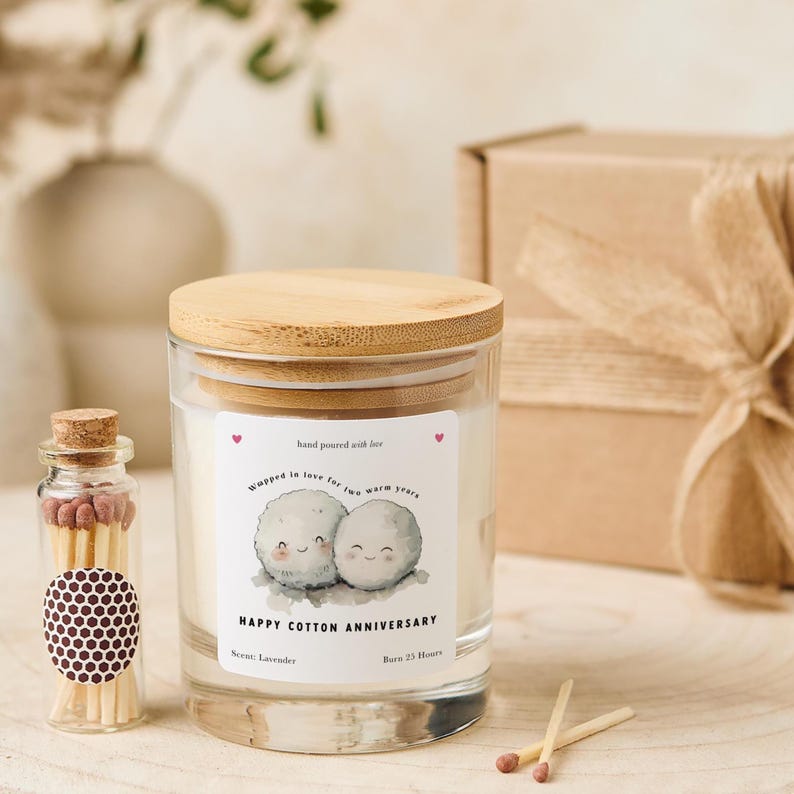 May include: A glass candle jar with a wooden lid, labeled "Happy Cotton Anniversary." The label features two cartoon cotton balls and the text "Wrapped in love for two warm years." A small glass bottle of matches and a gift box are also present.