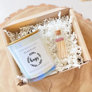 Peut inclure: Coffret cadeau contenant une bougie nacrée à bordure dorée et une petite bouteille d'allumettes. La bougie porte l'inscription "No words just hugs". La boîte est remplie de papier déchiqueté blanc.