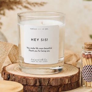 Pode incluir: Uma vela de cera branca num recipiente cilíndrico de vidro com um rótulo branco que apresenta o texto: VANILLA — 25 HRS. HEY SIS! You make life more beautiful. Thank you for being you. hand-poured with love VEGAN SOY WAX CANDLE.