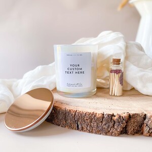 以下が含まれることがあります： ゴールドの蓋と「YOUR CUSTOM TEXT HERE」と「hand-poured with love, soy wax candle」と書かれたラベルが付いた白いキャンドル。キャンドルは木の切り株の上に置かれ、小さなガラス瓶に入ったマッチが添えられています。