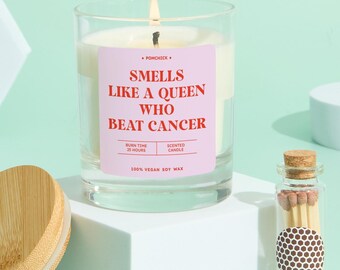May include: A lit scented candle in a clear glass jar with a pink label that reads "Smells Like a Queen Who Beat Cancer." The candle is made of 100% vegan soy wax and has a burn time of 25 hours. A small glass bottle of matches is next to the candle.