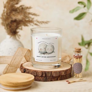 May include: A clear glass candle with a label that reads "Happy Cotton Anniversary" and features two cartoon cotton balls. The candle sits on a wooden slice with a wooden lid and a small glass jar of matches.