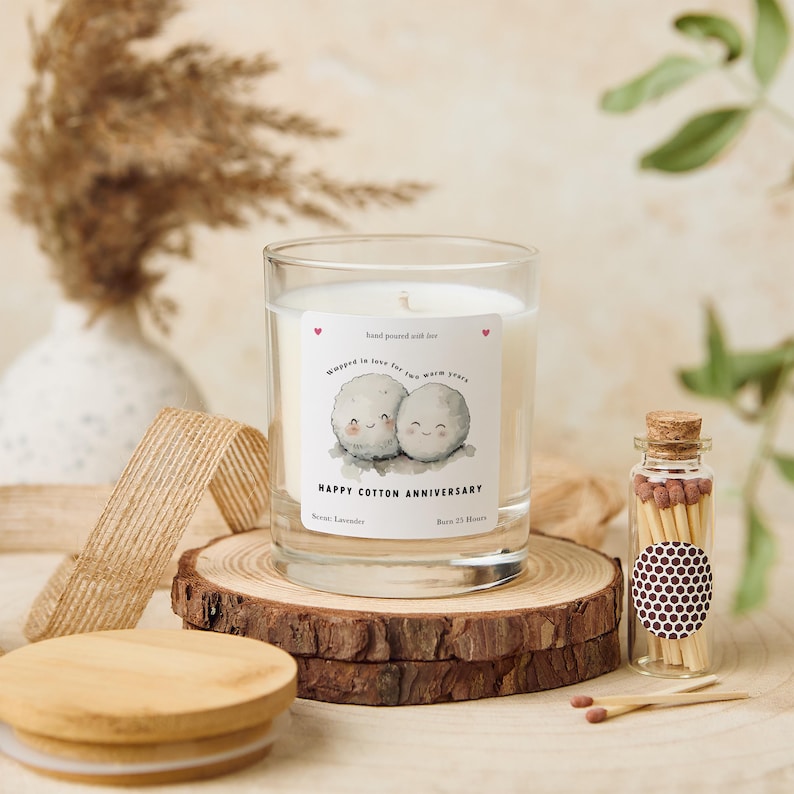 May include: A clear glass candle with a label that reads "Happy Cotton Anniversary" and features two cartoon cotton balls. The candle sits on a wooden slice with a wooden lid and a small glass jar of matches.