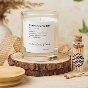 Op de afbeelding: Een heldere glazen kaars met de tekst "Forever starts here" en "Emily & Jack" gedrukt op een wit etiket. De kaars staat op een houten schijf met een klein glazen potje lucifers ernaast. De kaars is geurig naar lavendel.