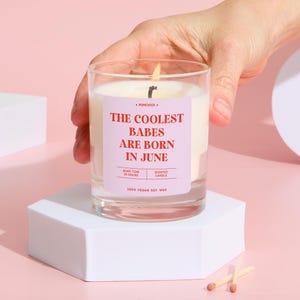 May include: A lit scented candle in a clear glass jar. The candle has a pink label with the text "THE COOLEST BABES ARE BORN IN JUNE". The candle is made of 100% vegan soy wax and has a burn time of 25 hours. The candle is on a white hexagonal stand.