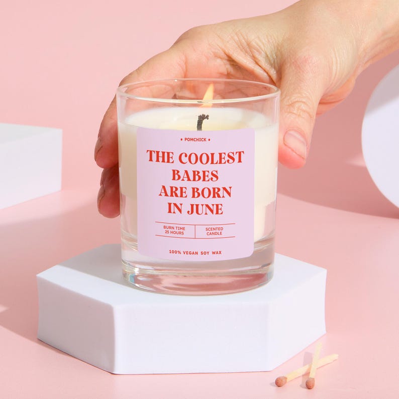 May include: A lit scented candle in a clear glass jar. The candle has a pink label with the text "THE COOLEST BABES ARE BORN IN JUNE". The candle is made of 100% vegan soy wax and has a burn time of 25 hours. The candle is on a white hexagonal stand.
