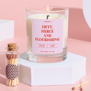 Pode incluir: Uma vela perfumada acesa em um frasco de vidro transparente com um rótulo rosa que diz "FIFTY, FIERCE AND FLOURISHING". Uma pequena garrafa de vidro de fósforos fica ao lado da vela. A vela é feita de cera de soja 100% vegana e tem um tempo de queima de 25 horas.