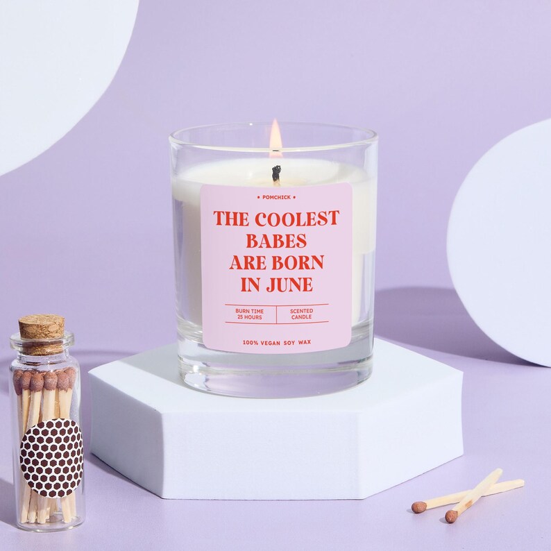 May include: A lit scented candle in a clear glass jar with a pink label that reads "THE COOLEST BABES ARE BORN IN JUNE." The candle sits on a white hexagonal stand. A small glass bottle of matches is next to the candle.