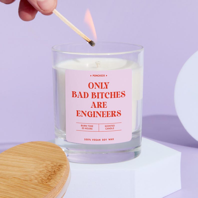 May include: A clear glass candle with a pink label that reads "ONLY BAD BITCHES ARE ENGINEERS." A lit match is held above the candle. The candle is on a white surface against a purple background. The candle is made of 100% vegan soy wax.